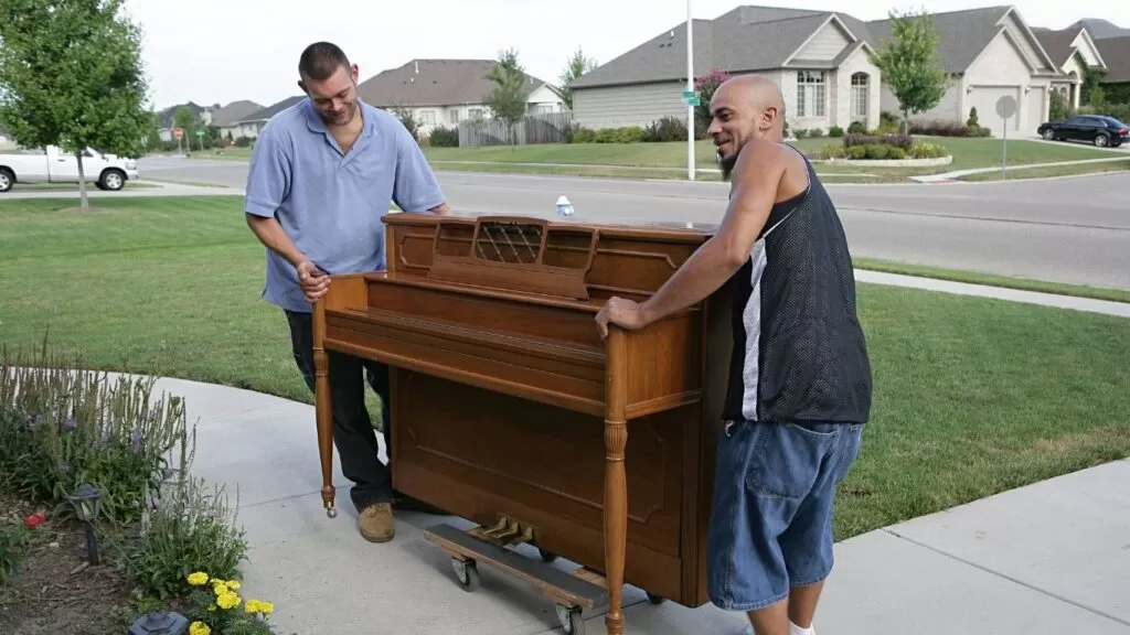 Dois homens guiando piano que esta encima de uma prancha com rodas.